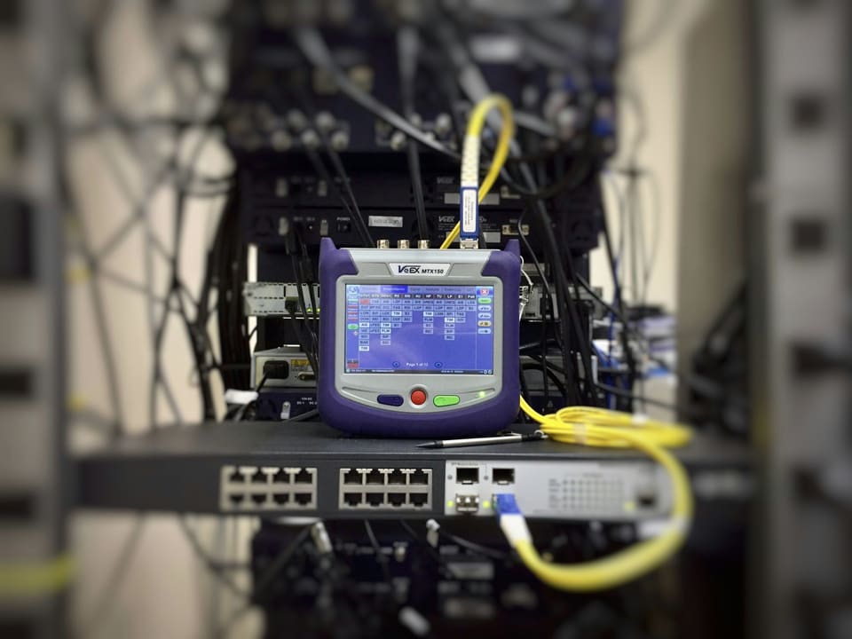 A handheld network tester connected by fibre and copper leads to a switch in a rack.