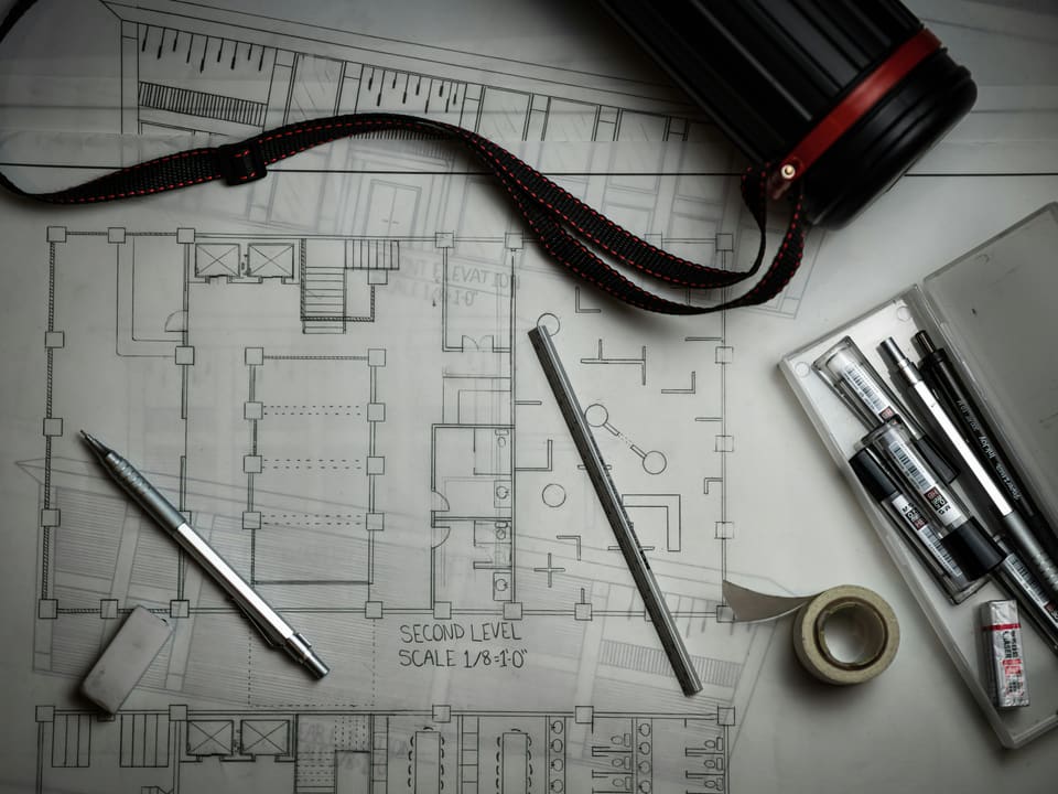 Architectural floor plans on a desk with drafting pencils, a scale rule and tape.